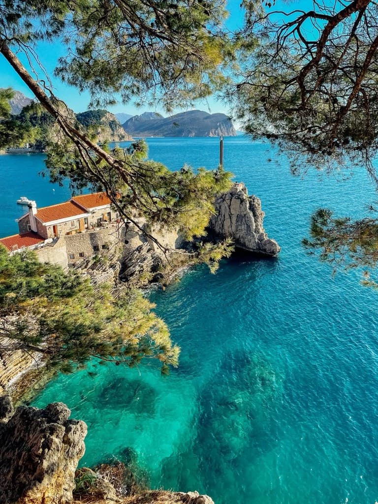 Turquoise Adriatic Sea below a stone house on a rocky cliff, framed by pine branches, Montenegro coast