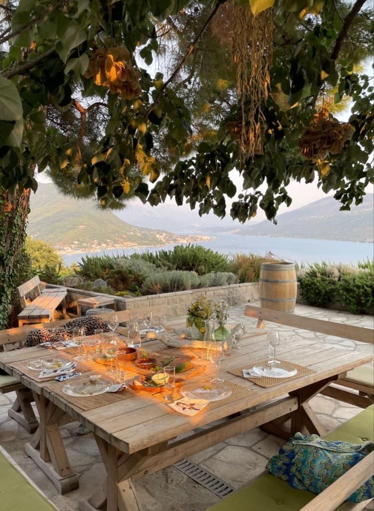 Outdoor wine and dining on a stone terrace at sunset, table set with glasses and platters, bay and mountains in the distance, Montenegro